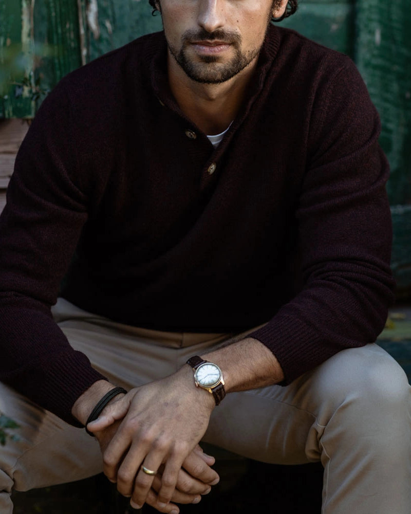 A man wearing a dark burgundy knit henley sweater and beige trousers, sitting in front of a rustic green wooden door. He is wearing a brown leather watch with a white dial on his wrist.