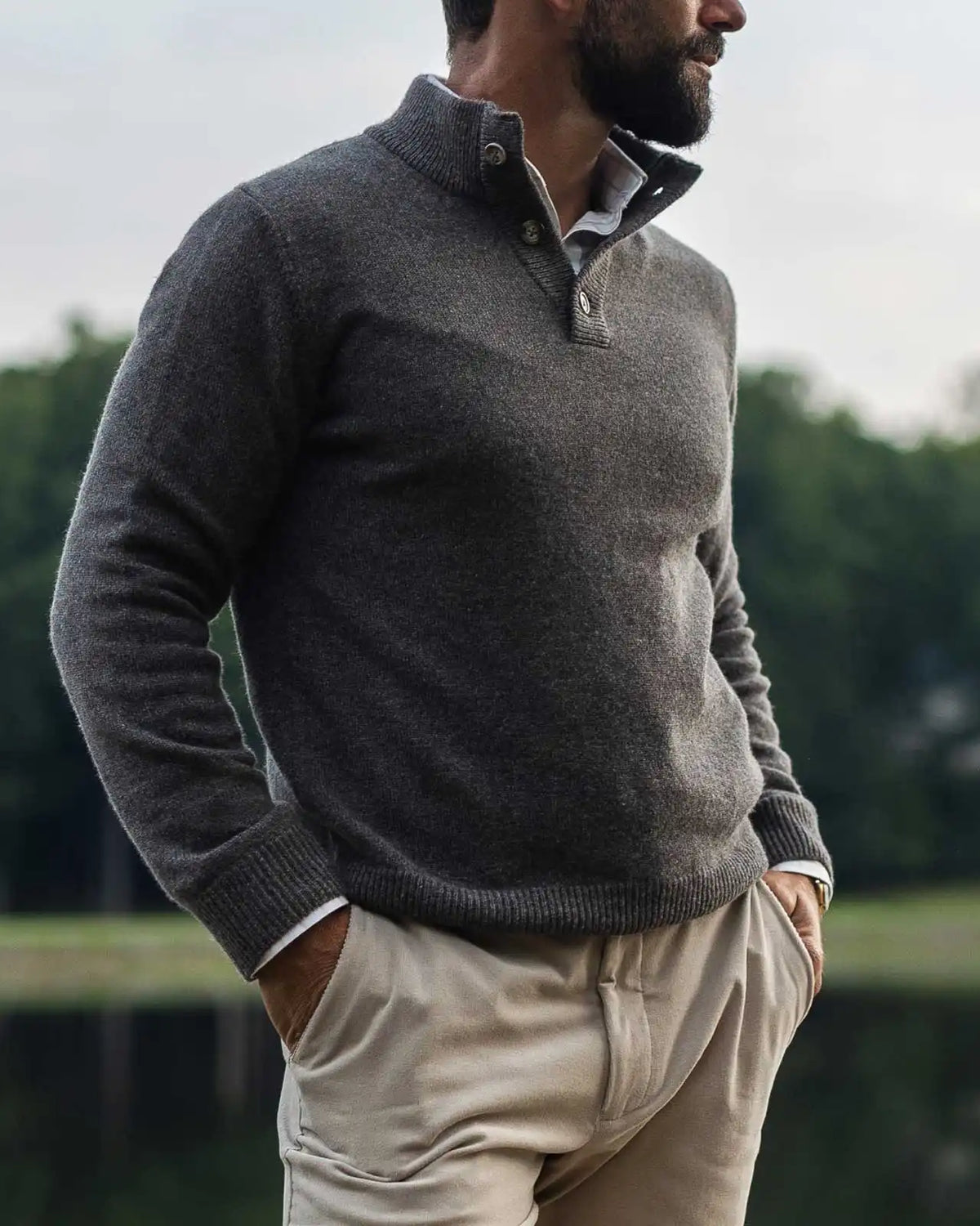 Man wearing a taupe light brown mock neck Italian merino sweater. He is standing outside in front of a lake with a blurred background.