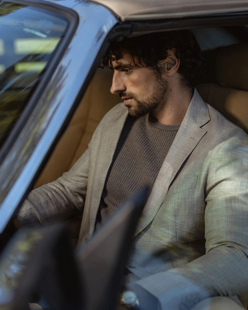 A man with dark curly hair and a beard sits in the driver's seat of a car, wearing a light beige blazer over a brown knit sweater.