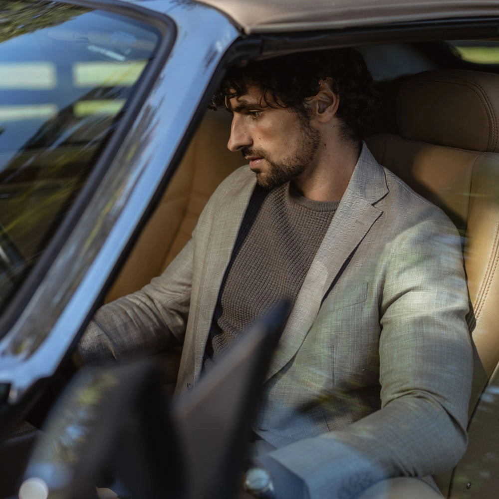 A man with dark curly hair and a beard sits in the driver's seat of a car, wearing a light beige blazer over a brown knit sweater.