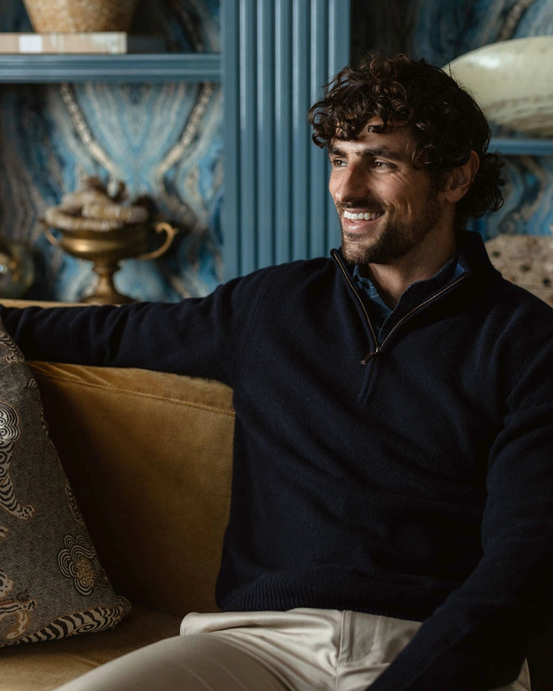 A smiling man with curly dark hair sits on a tan sofa wearing a dark navy quarter-zip sweater and light beige trousers, with a decorative pillow beside him in an ornate blue and gold room.