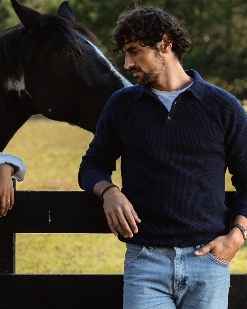 A man wearing a navy blue knit polo sweater and light blue jeans stands outdoors next to a black fence, with a horse visible behind him. The product being shown is the navy sweater.