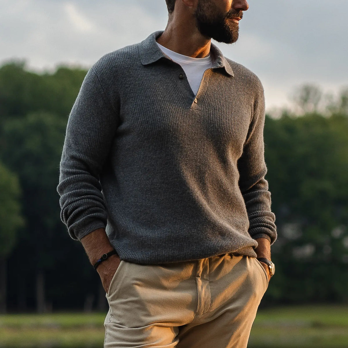 A man wearing a gray ribbed knit polo-neck sweater over a white t-shirt and khaki pants, standing outdoors with trees in the background.