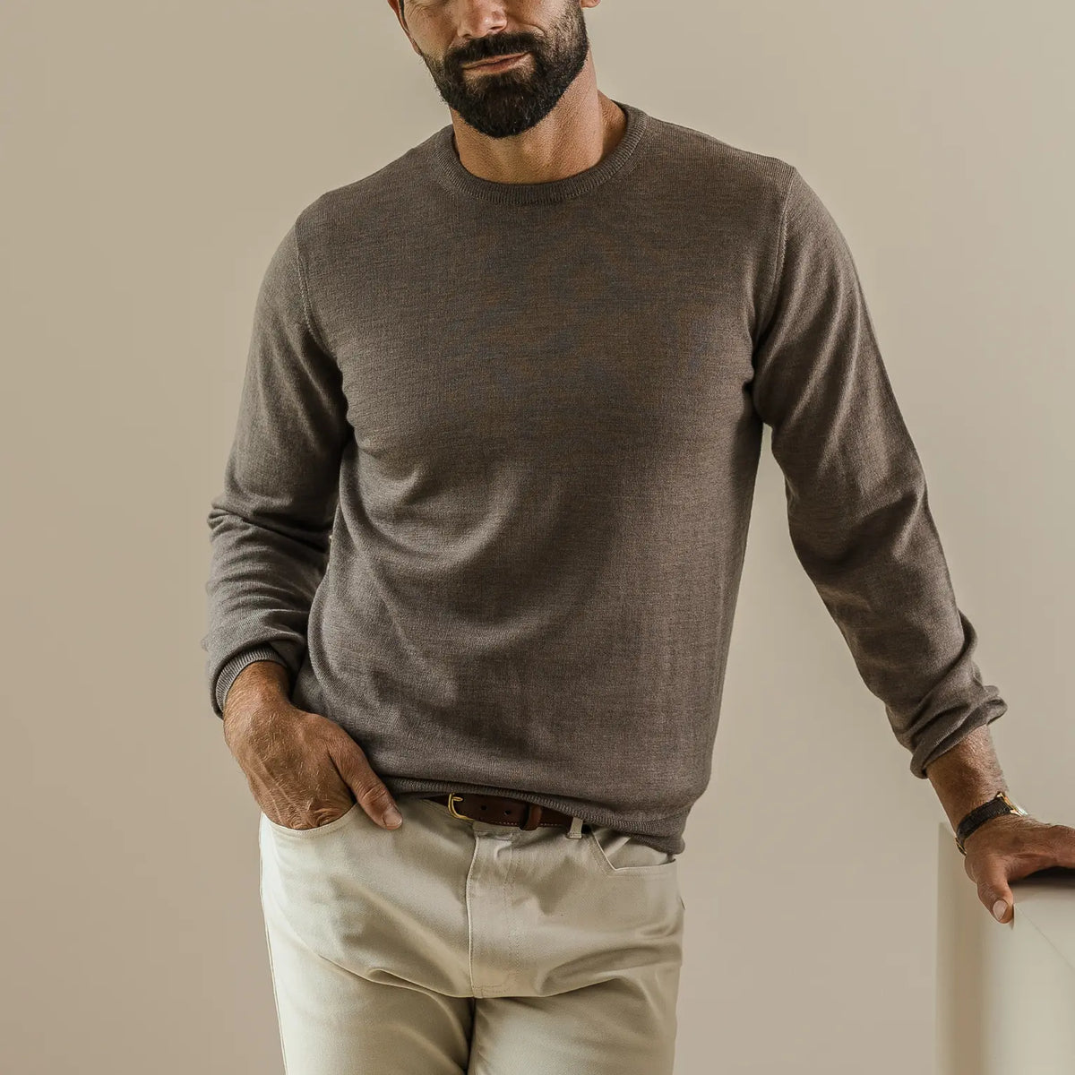A man wearing a light brown long-sleeve crewneck knit sweater and light beige trousers, standing against a neutral beige background.