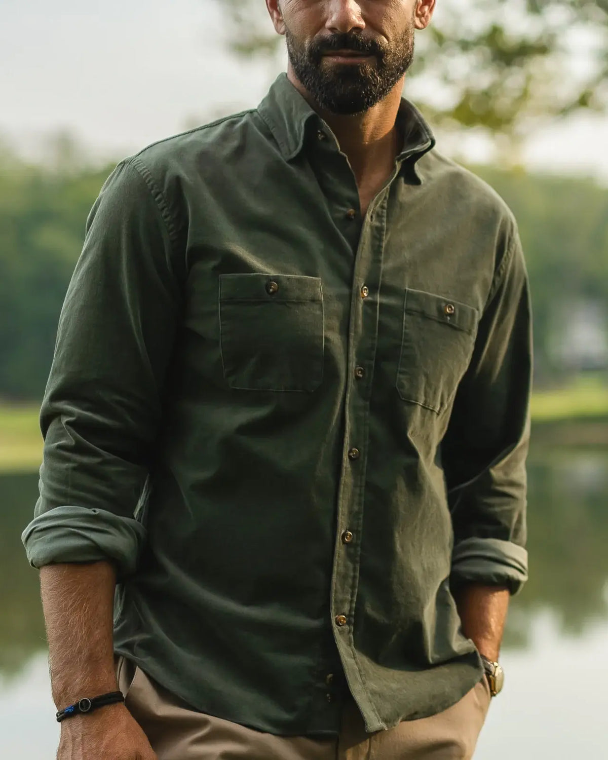 A man wearing an olive green long-sleeve button-up shirt with two chest pockets, standing outdoors near a body of water with trees in the background.