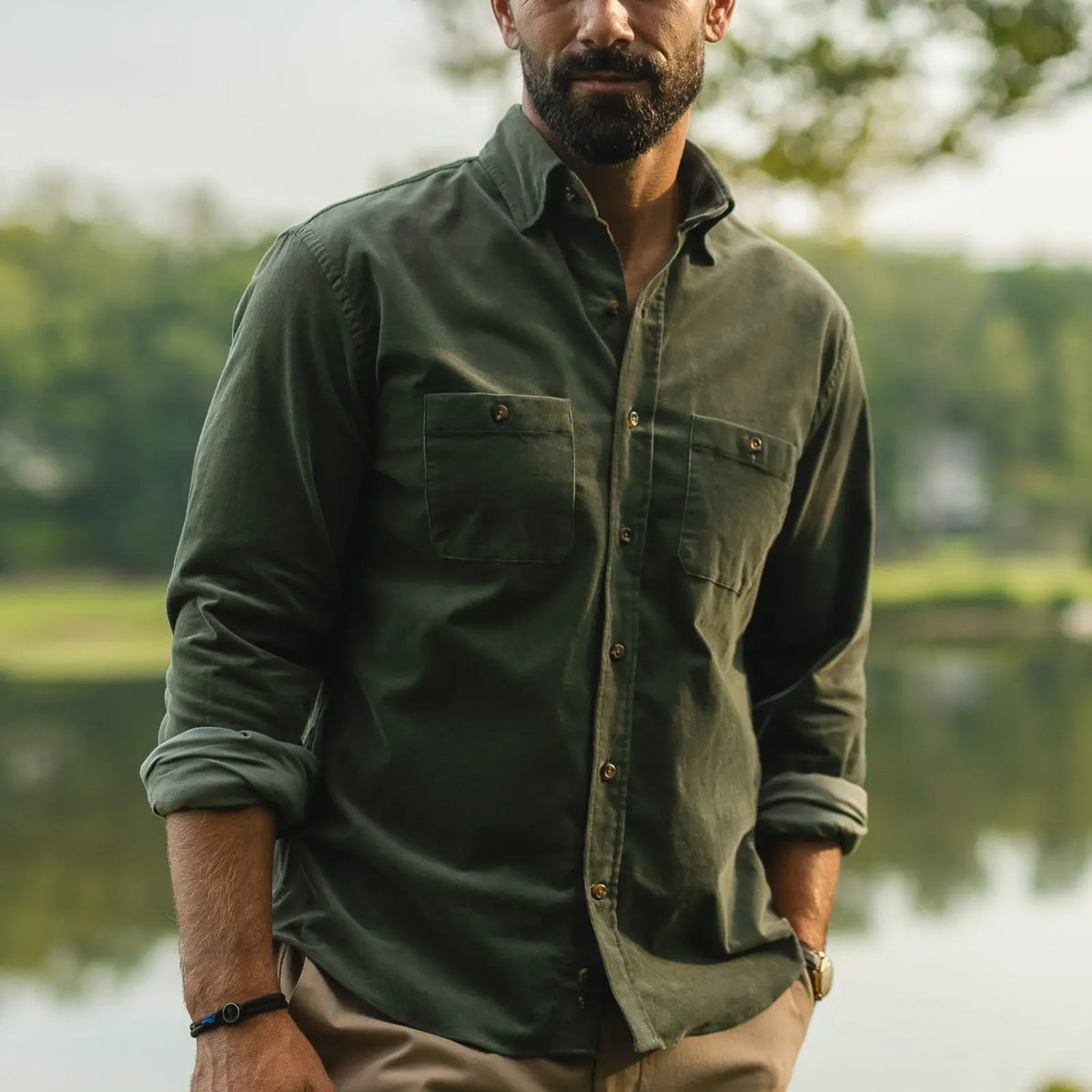 A man wearing an olive green long-sleeve button-up shirt with two chest pockets, standing outdoors near a body of water with trees in the background.