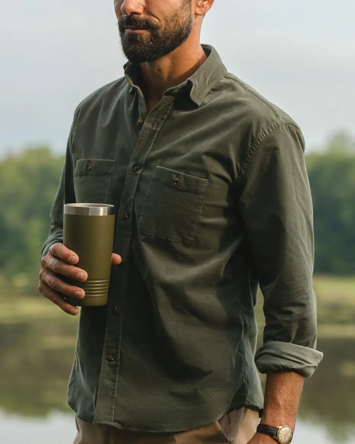 A man outdoors by a lake wearing an olive green long-sleeve button-up shirt and holding an olive green insulated tumbler.