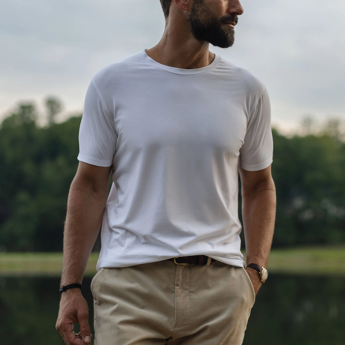 A man wearing a plain white short-sleeve crewneck t-shirt and khaki pants, standing outdoors with a blurred background of trees and water.
