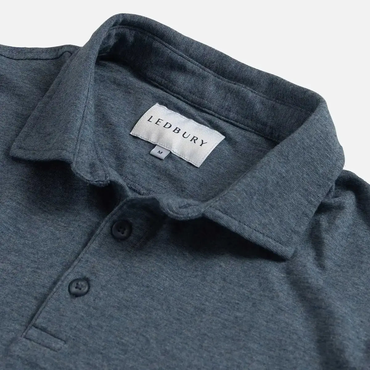 Close-up of a slate blue polo shirt collar with a visible LEDBURY neck label and two dark buttons on a light gray background.
