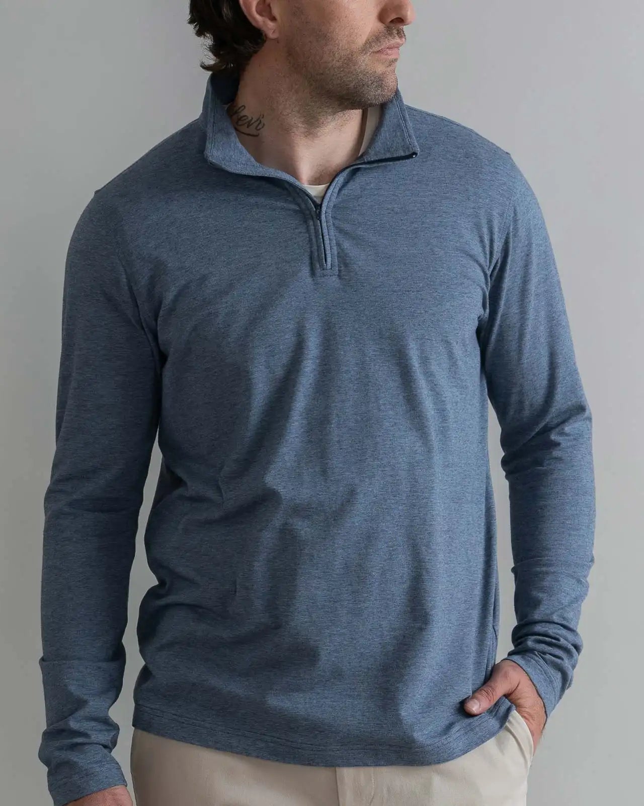 A man wearing a heathered slate blue long-sleeve quarter-zip pullover sweater, paired with beige pants, standing against a neutral gray background.