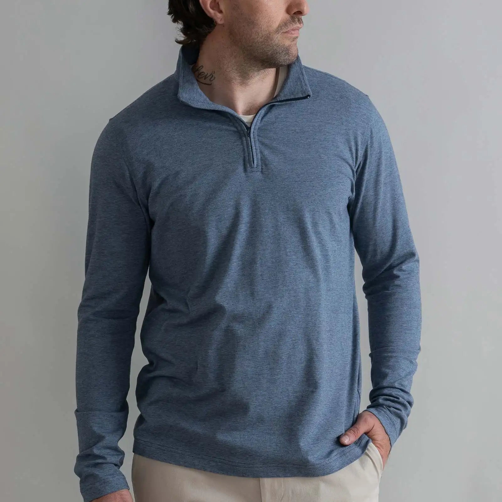 A man wearing a heathered slate blue long-sleeve quarter-zip pullover sweater, paired with beige pants, standing against a neutral gray background.