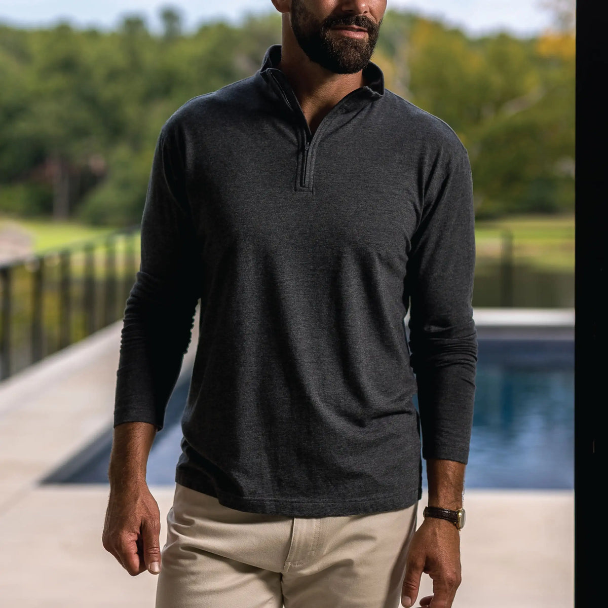 A man wearing a dark charcoal gray long-sleeve quarter-zip pullover shirt and beige pants, standing outdoors near a pool with a blurred green background.