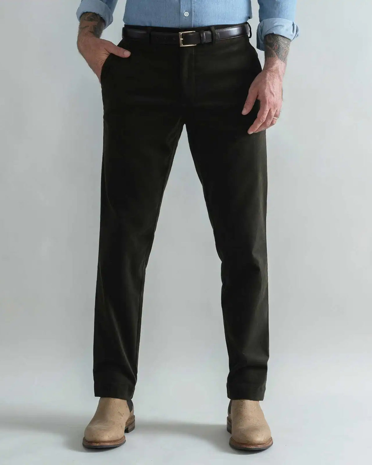 A man wearing slim-fit dark green corduroy trousers, a black belt, a light blue button-down shirt, and tan boots, standing against a light gray studio background.
