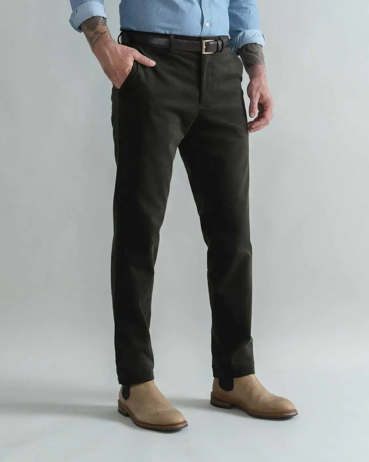 A man wearing slim-fit dark olive green corduroy trousers, styled with a black belt, a light blue shirt, and tan suede boots, standing against a light gray studio background.