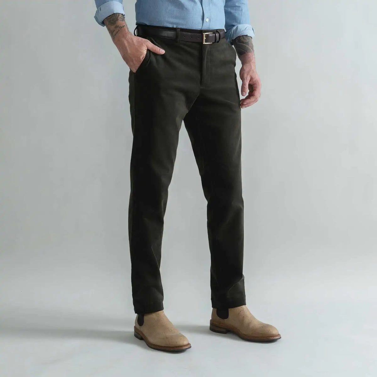 A man wearing slim-fit dark olive green corduroy trousers, styled with a black belt, a light blue shirt, and tan suede boots, standing against a light gray studio background.