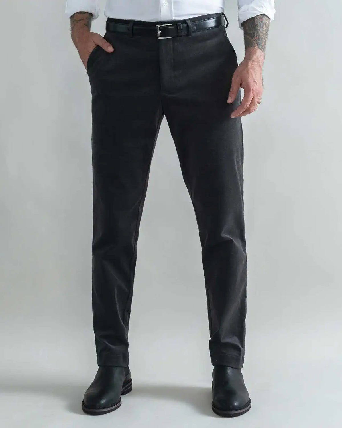 A man wearing slim-fit dark grey corduroy trousers, a black belt, a white shirt, and black leather boots, standing against a light gray studio background.