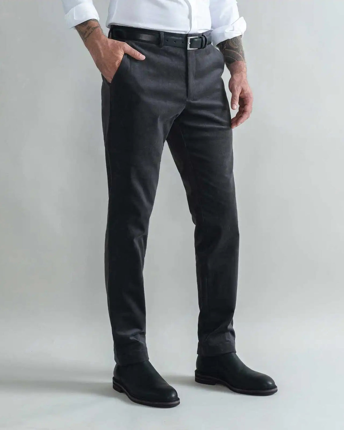 A man wearing slim-fit charcoal gray corduroy chinos, styled with a white shirt, black belt, and black boots, standing against a light gray studio background.