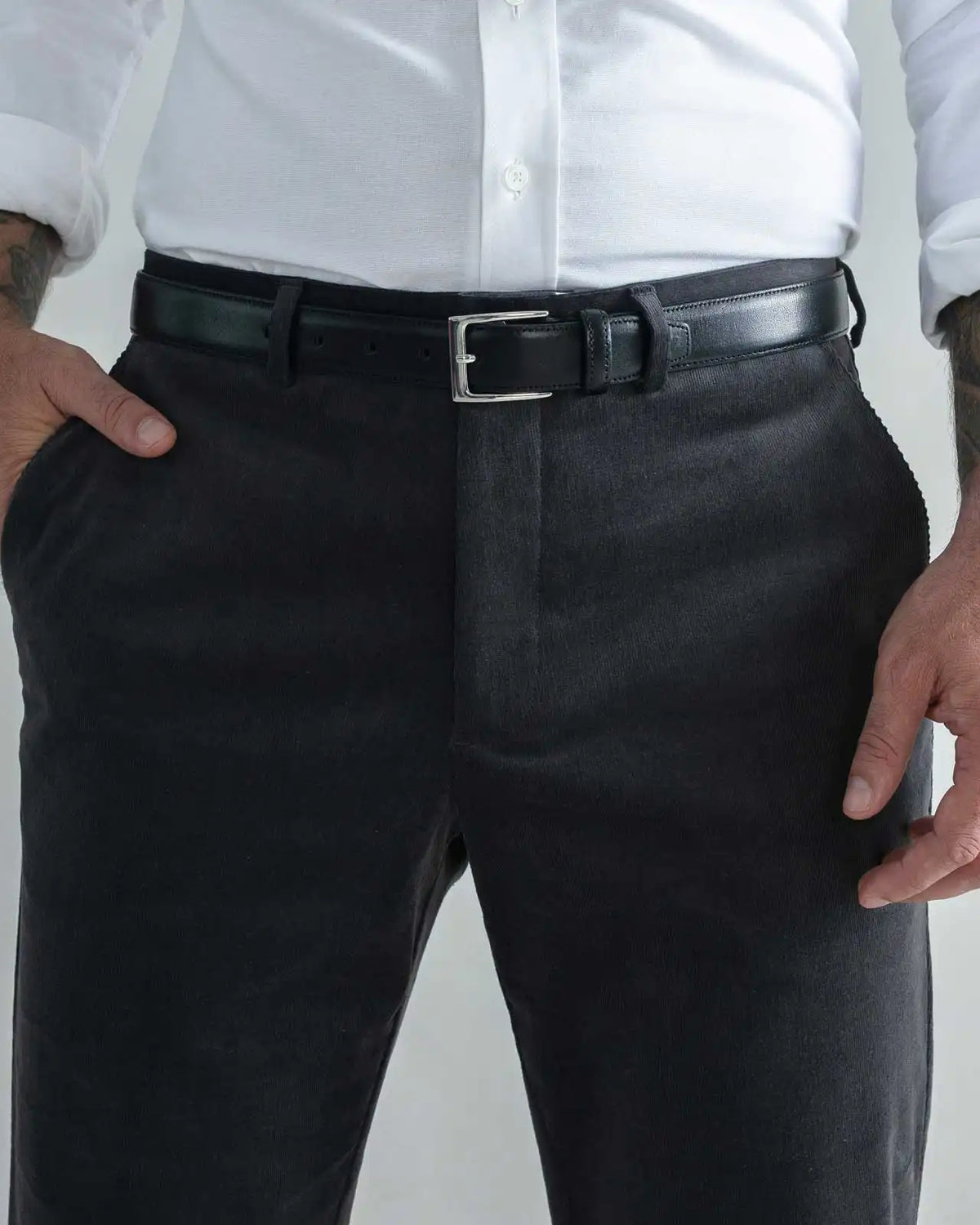 A close-up of a man wearing dark charcoal gray corduroy trousers, a black leather belt with a silver buckle, a white button-down shirt, and tattoos on both arms, set against a plain light gray background.