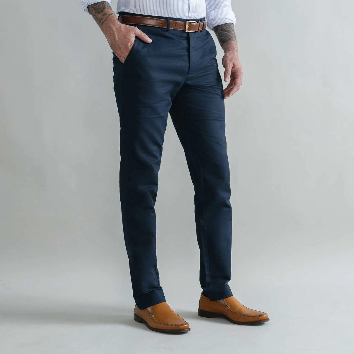 A man wearing slim-fit navy blue trousers, styled with a brown leather belt, a white shirt, and tan leather shoes, standing against a light gray studio background.