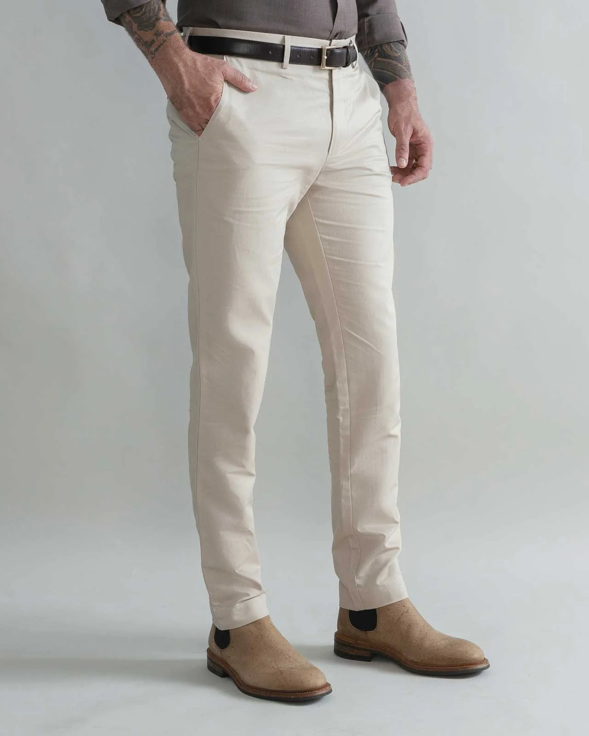 A man wearing slim-fit khaki trousers, styled with a black belt, a gray shirt, and tan suede Chelsea boots, shown from the waist down against a light gray studio background.