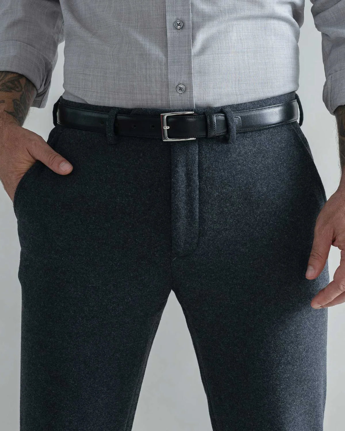 A man wearing dark charcoal gray wool trousers, a light gray button-down shirt, and a black leather belt with a silver buckle, shown from the waist down against a light gray background.
