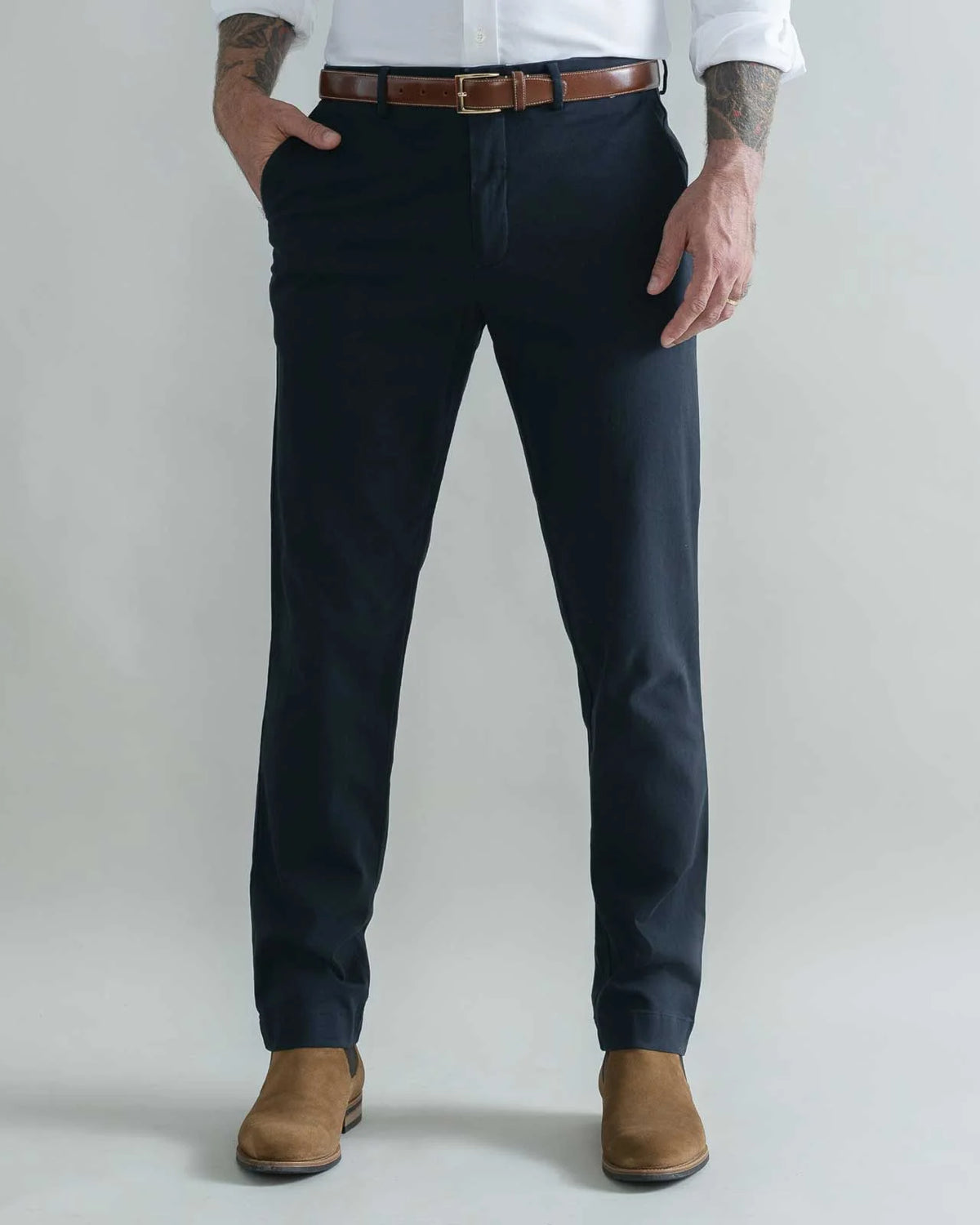 A man wearing dark navy blue slim-fit trousers, styled with a brown leather belt, a white dress shirt, and brown suede boots, standing against a light gray studio background.