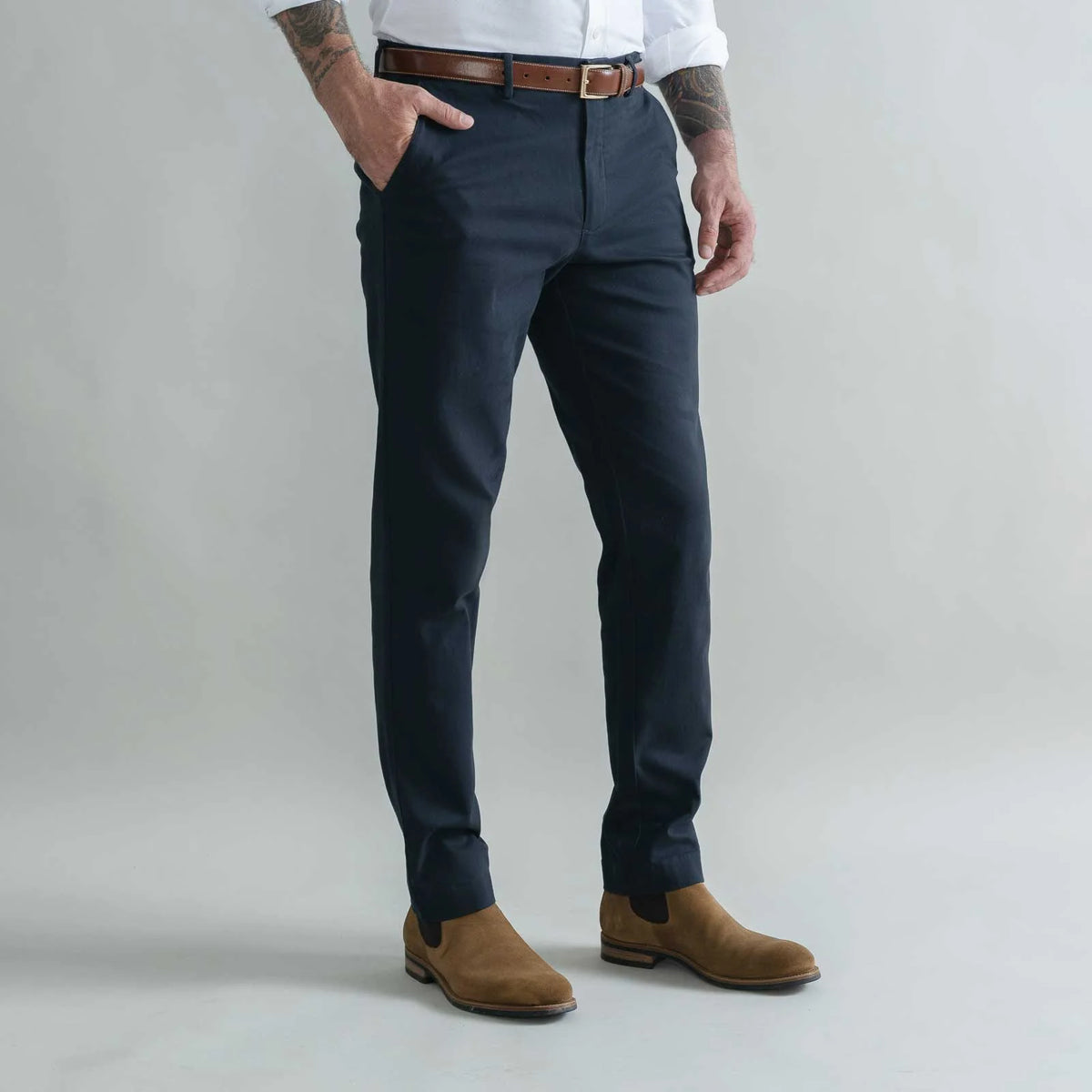 A man wearing navy blue slim-fit trousers, styled with a brown leather belt, a white shirt, and brown suede Chelsea boots, standing against a light gray studio background.