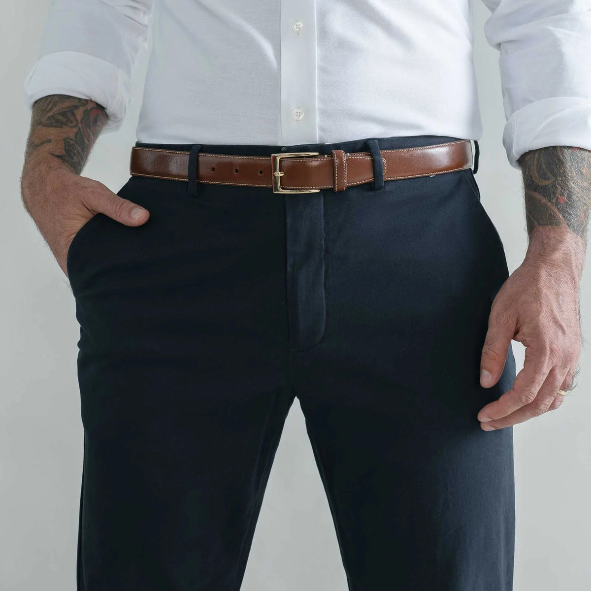 A man wearing dark navy trousers, a brown leather belt with a gold buckle, a white button-down shirt, and tattoos on both arms, standing against a light gray studio background.