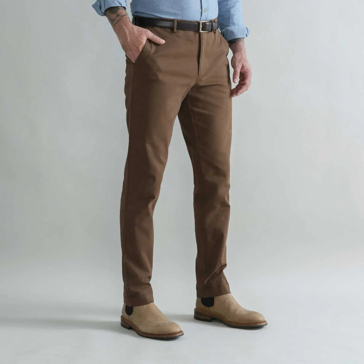 A man wearing slim-fit brown trousers, a dark brown belt, a light blue shirt, and tan suede boots, standing against a light gray studio background.