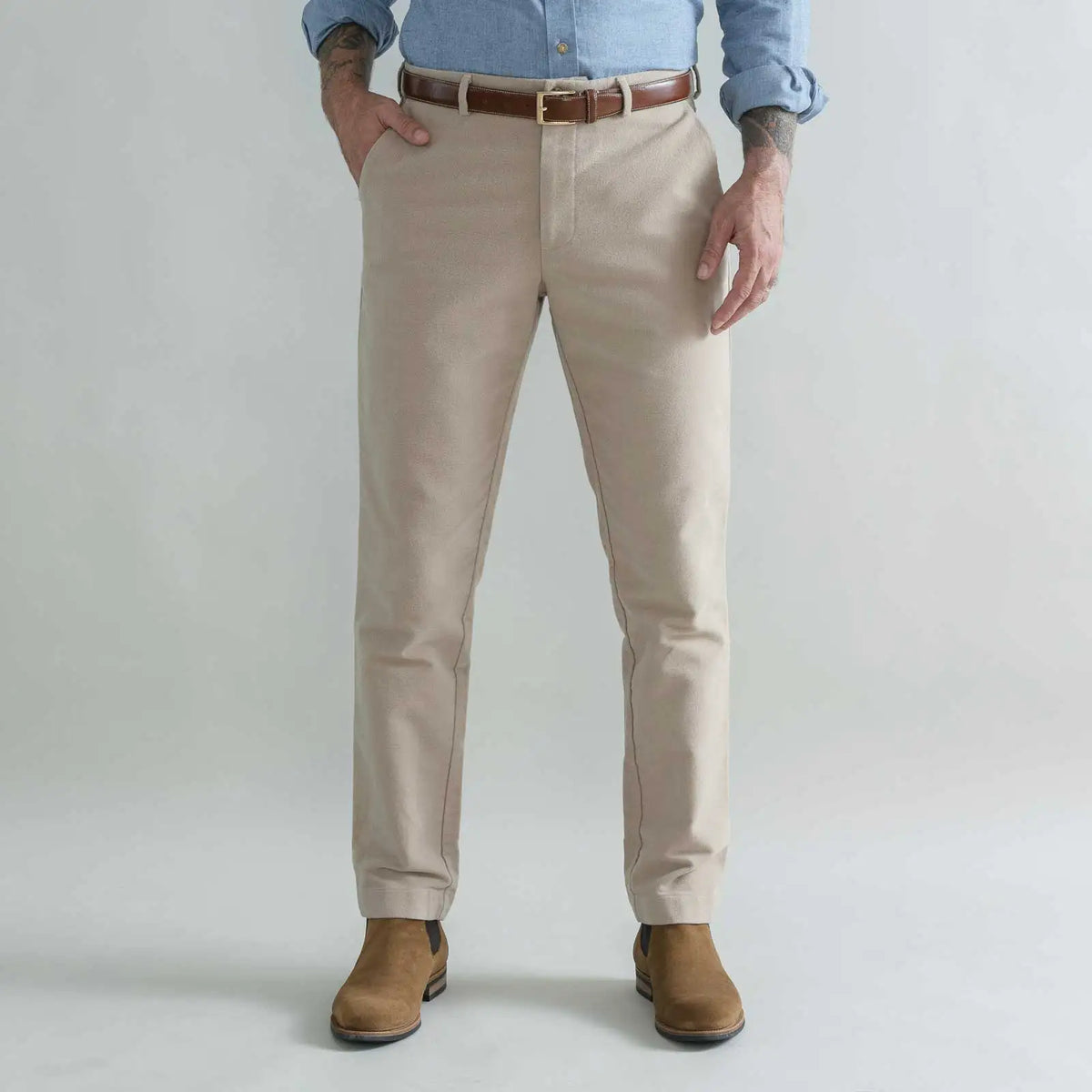 A man wearing tan moleskin chino pants with a brown belt, a light blue button-down shirt, and brown boots, standing against a light gray studio background.