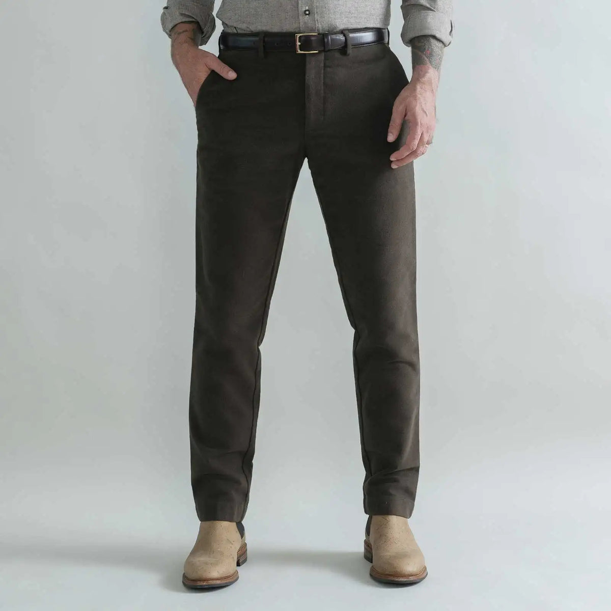 A person wearing slim-fit dark brown moleskin trousers, styled with a black belt, a gray shirt, and beige boots, standing against a light gray studio background