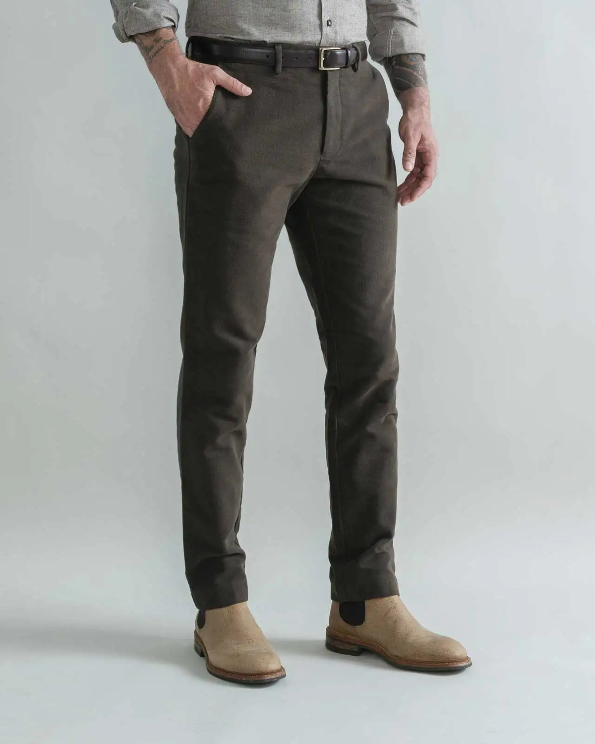 A man wearing slim-fit dark brown moleskin trousers, a gray shirt, and tan Chelsea boots, standing against a light gray studio background.