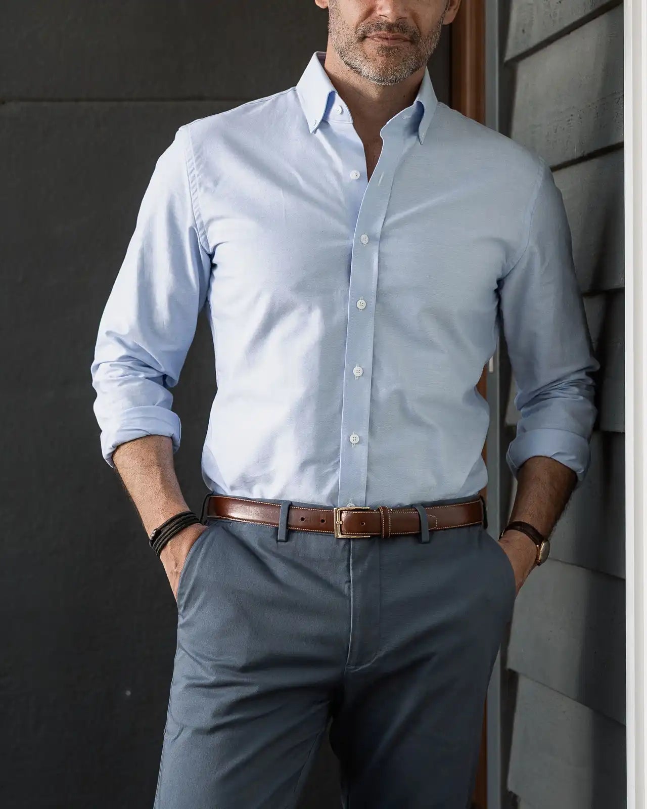 Man wearing a light blue Oxford dress shirt and gray pants against a dark wall.