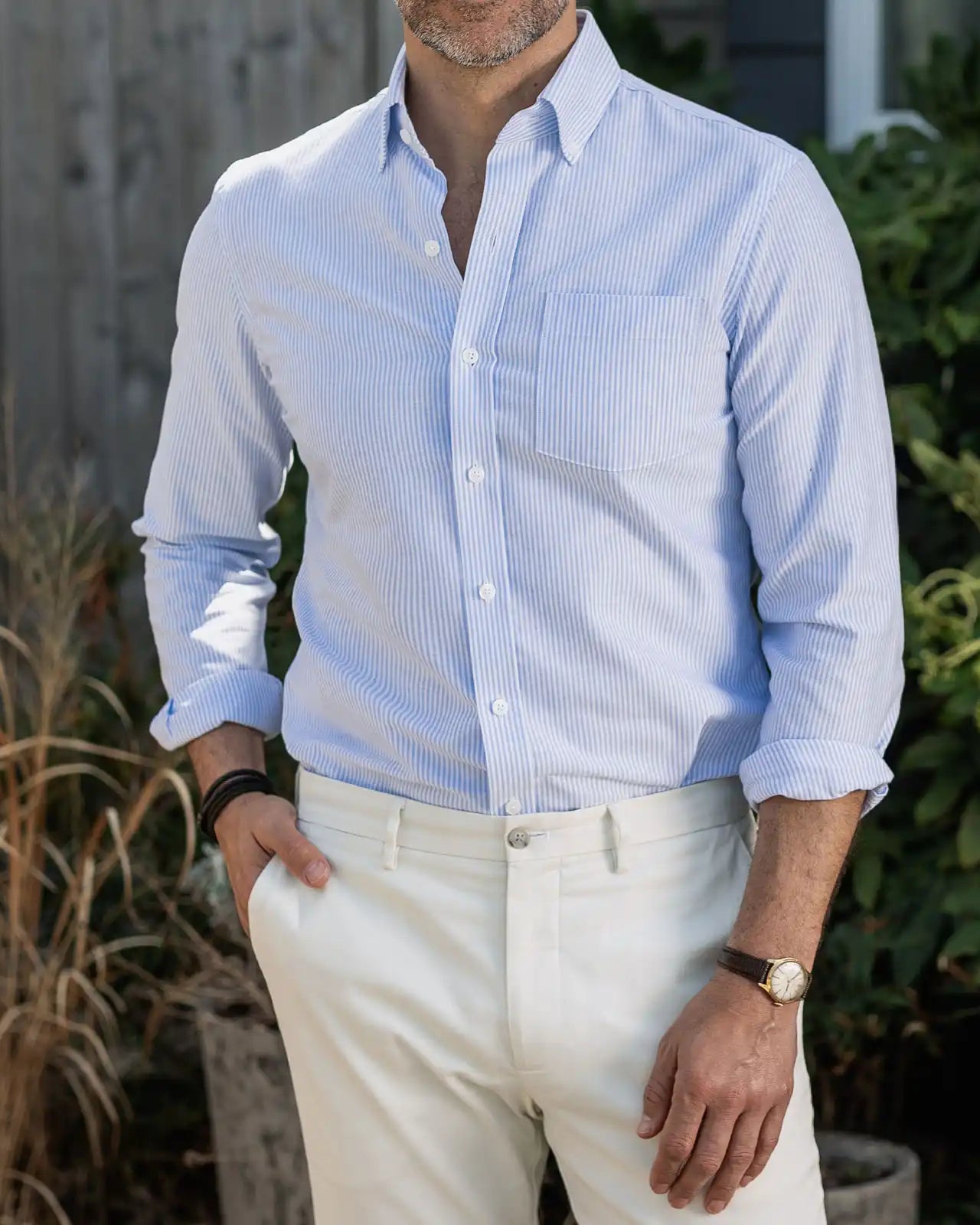 Man wearing a light blue striped Mayfield Oxford shirt and white pants standing outdoors.