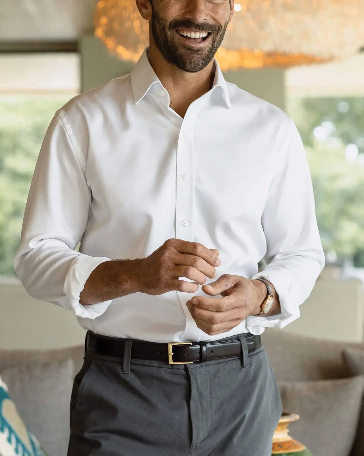 A man wearing a crisp white long-sleeve button-down dress shirt, dark gray trousers, and a black belt with a gold buckle, standing in a modern living room setting.