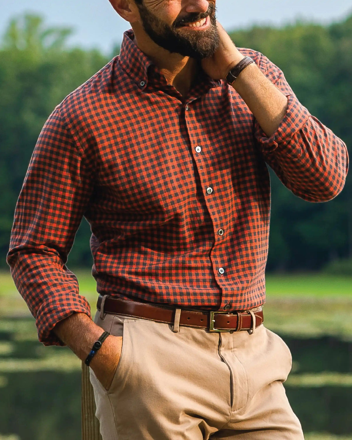 A man wearing an orange and black checkered long-sleeve button-down shirt, khaki pants, and a brown belt, standing outdoors in front of a blurred green background.