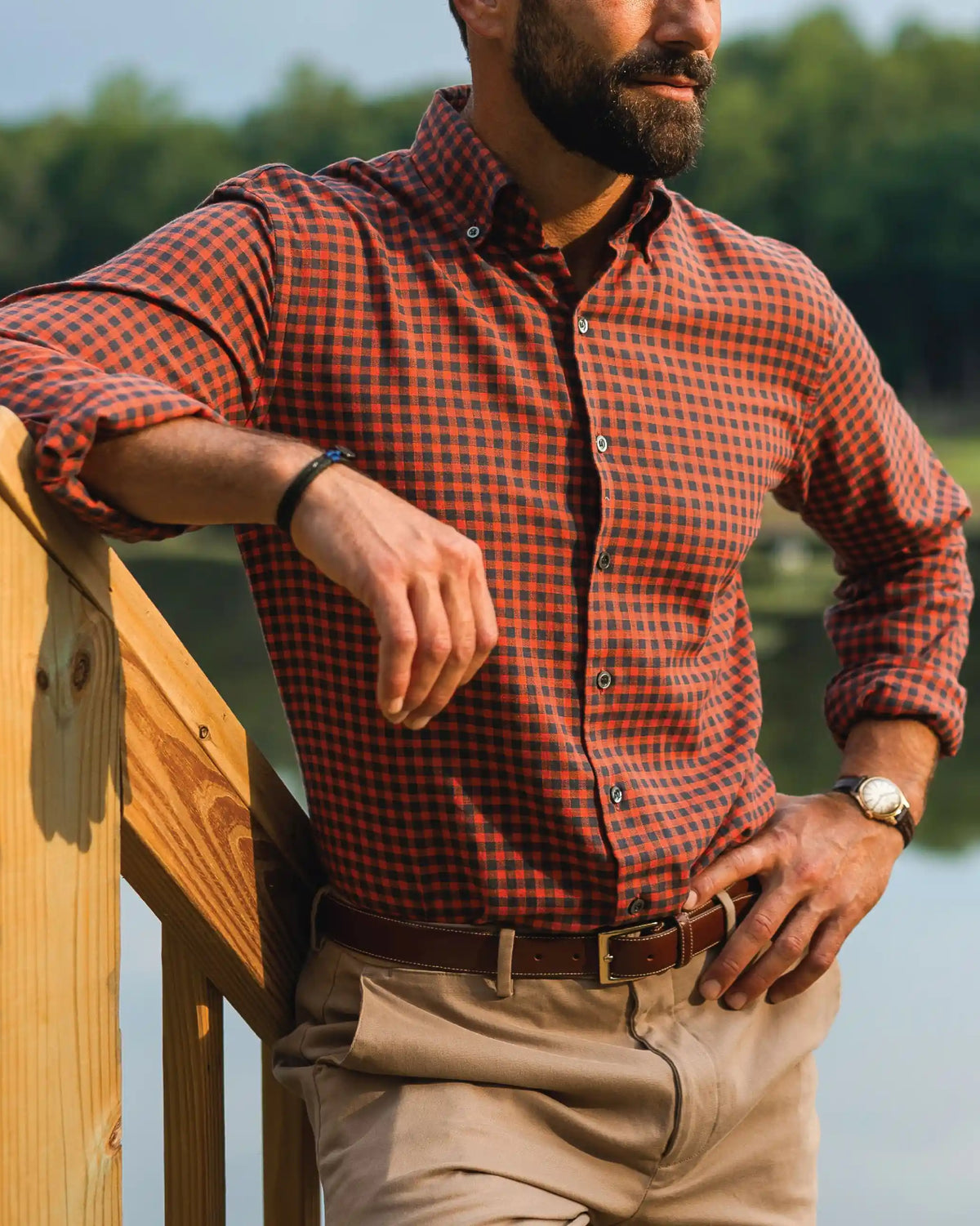A man wearing an orange and black checkered long-sleeve button-down shirt, khaki pants, and a brown leather belt, leaning on a wooden railing outdoors near a lake.