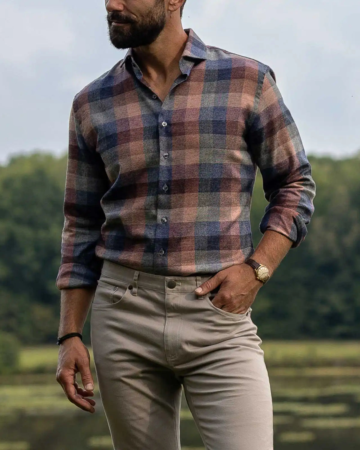 A man wearing a long-sleeve button-up plaid shirt in muted blue, red, and brown tones, paired with beige trousers, standing outdoors with trees in the background.