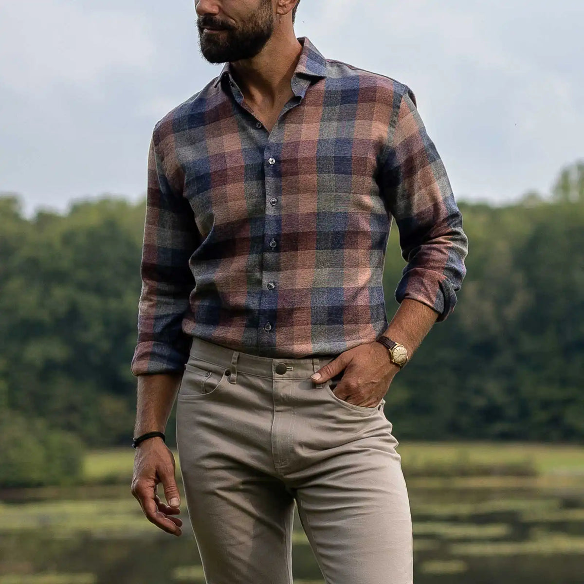 A man wearing a long-sleeve button-up plaid shirt in muted blue, red, and brown tones, paired with beige trousers, standing outdoors with trees in the background.