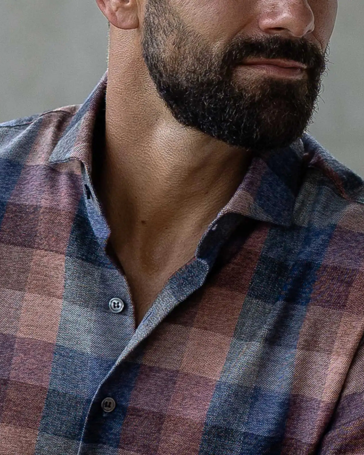 A close up of a plaid shirt in muted blues and orange tones on a man with a beard looking off to the side, with his eyes cut off just above the nose.
