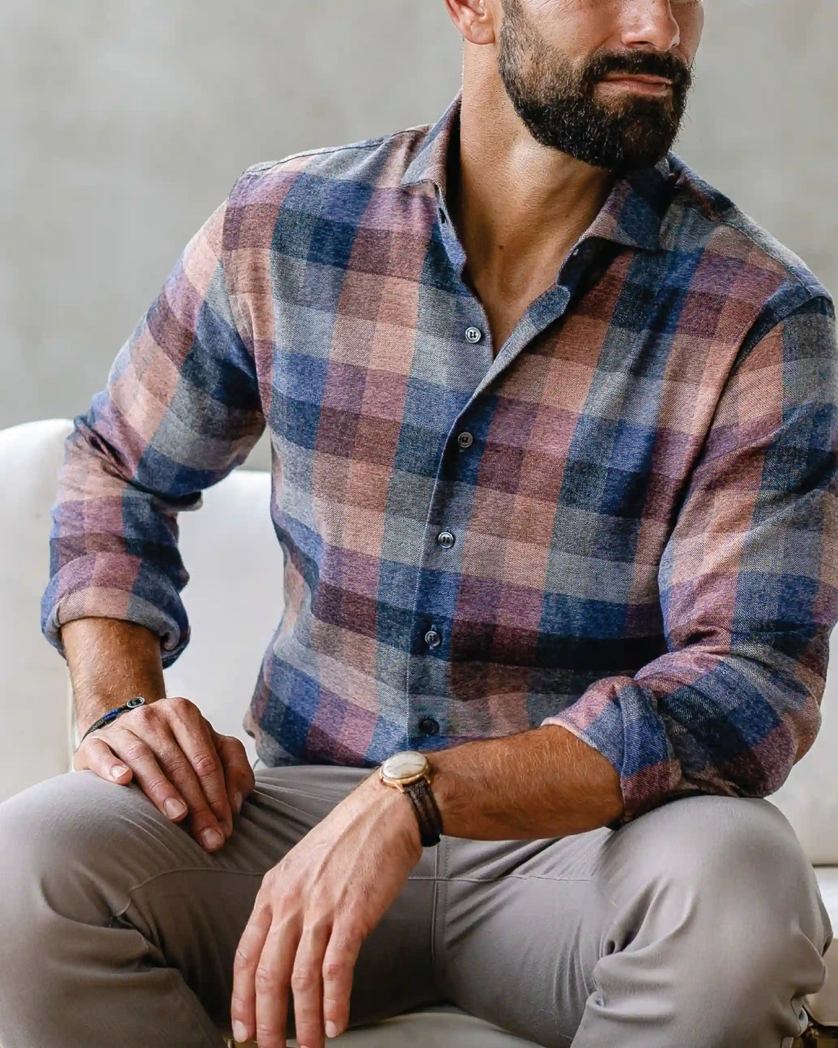 A man wearing a long-sleeve button-down plaid shirt in muted blue, pink, and beige tones, paired with light gray trousers, sitting on a white couch.