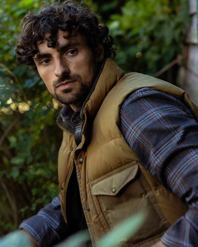 A man wearing a tan quilted puffer vest over a dark blue and gray plaid flannel shirt, photographed outdoors against a blurred green garden background.