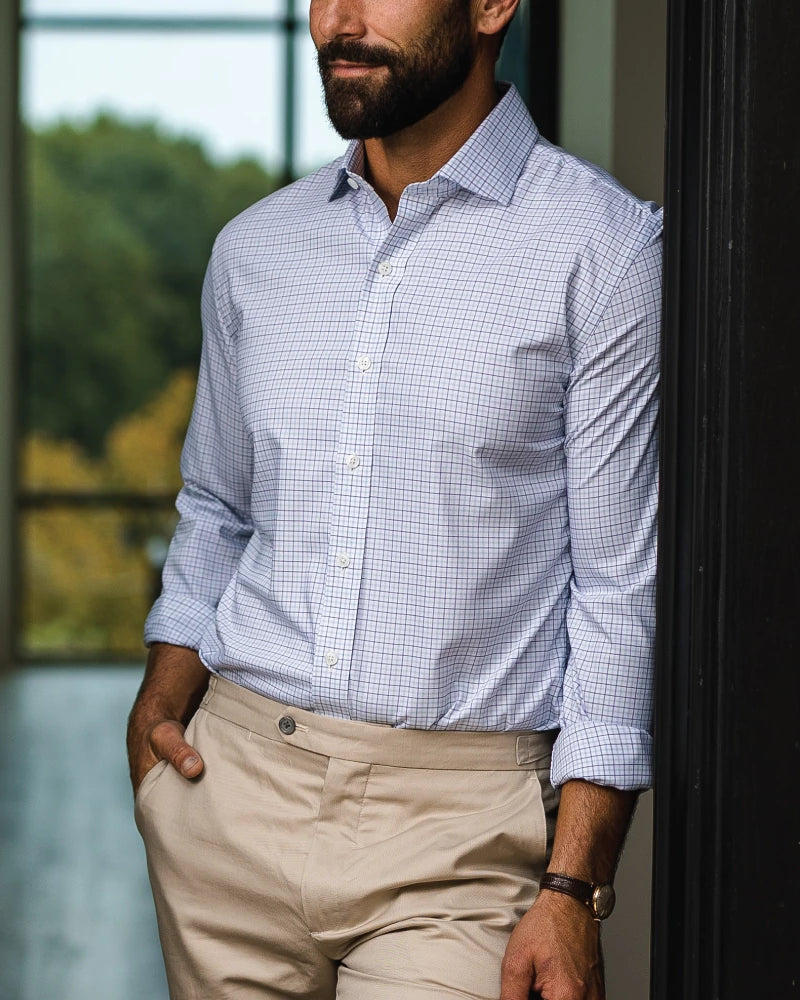 A man wearing a light blue and white checkered long-sleeve button-down shirt tucked into beige trousers, standing indoors near a window.