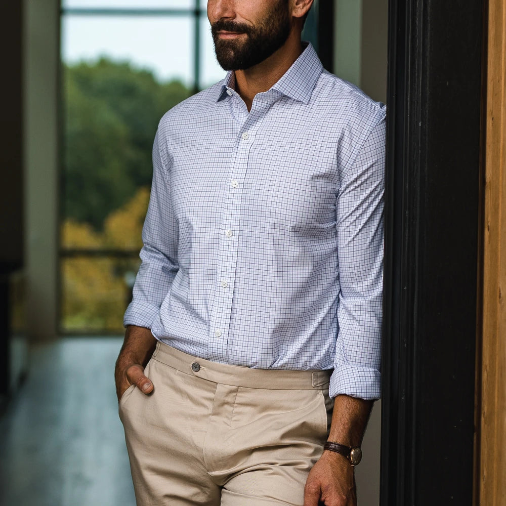 A man wearing a light blue and white checkered long-sleeve button-down shirt tucked into beige trousers, standing indoors near a window.