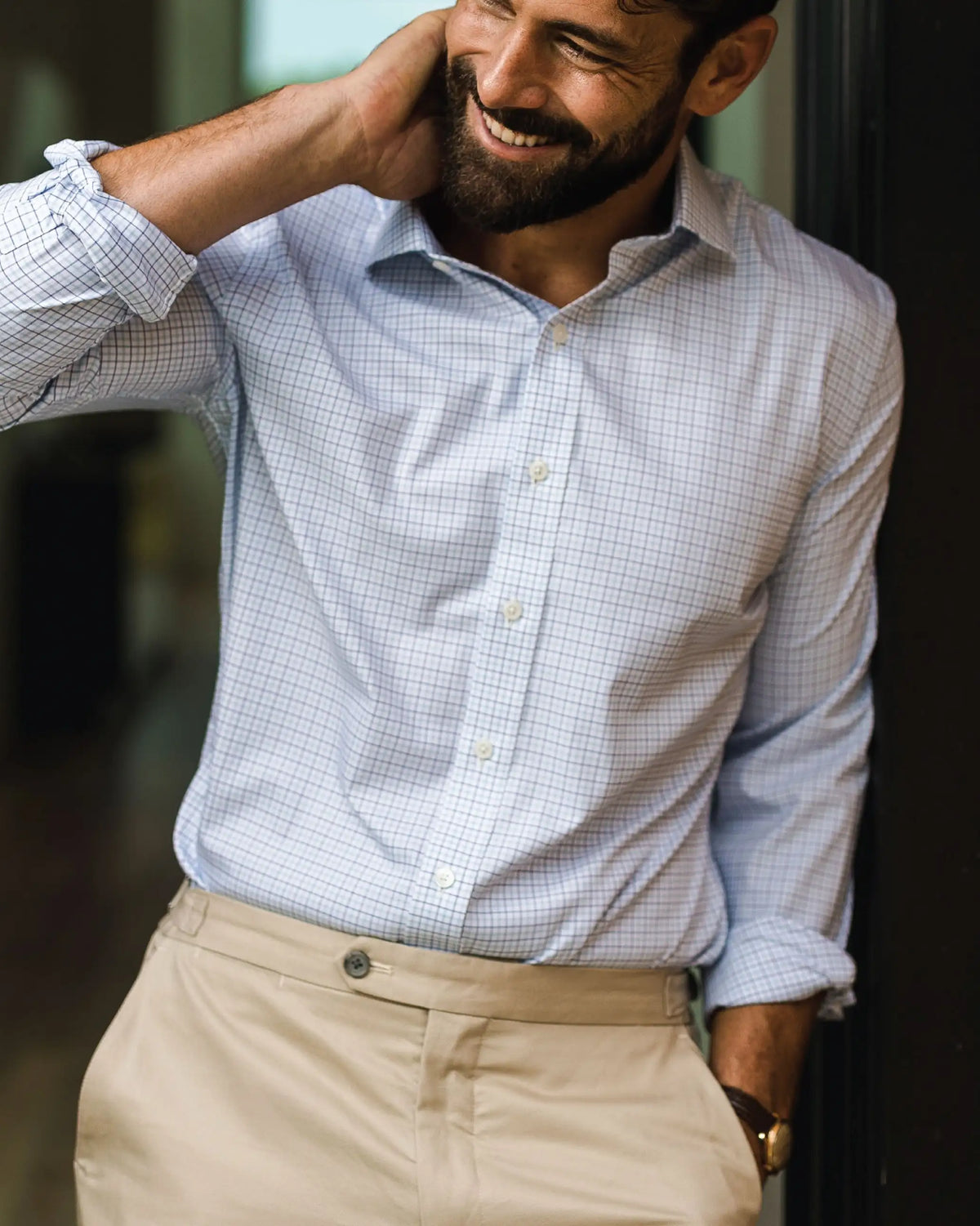 A man wearing a light blue and white checkered long-sleeve button-down shirt tucked into beige trousers, smiling and resting his head on his hand.