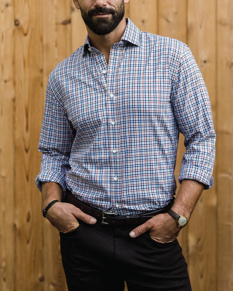 A man wearing a blue, white, and dark brown plaid button-down shirt with black trousers, standing in front of a wooden fence.