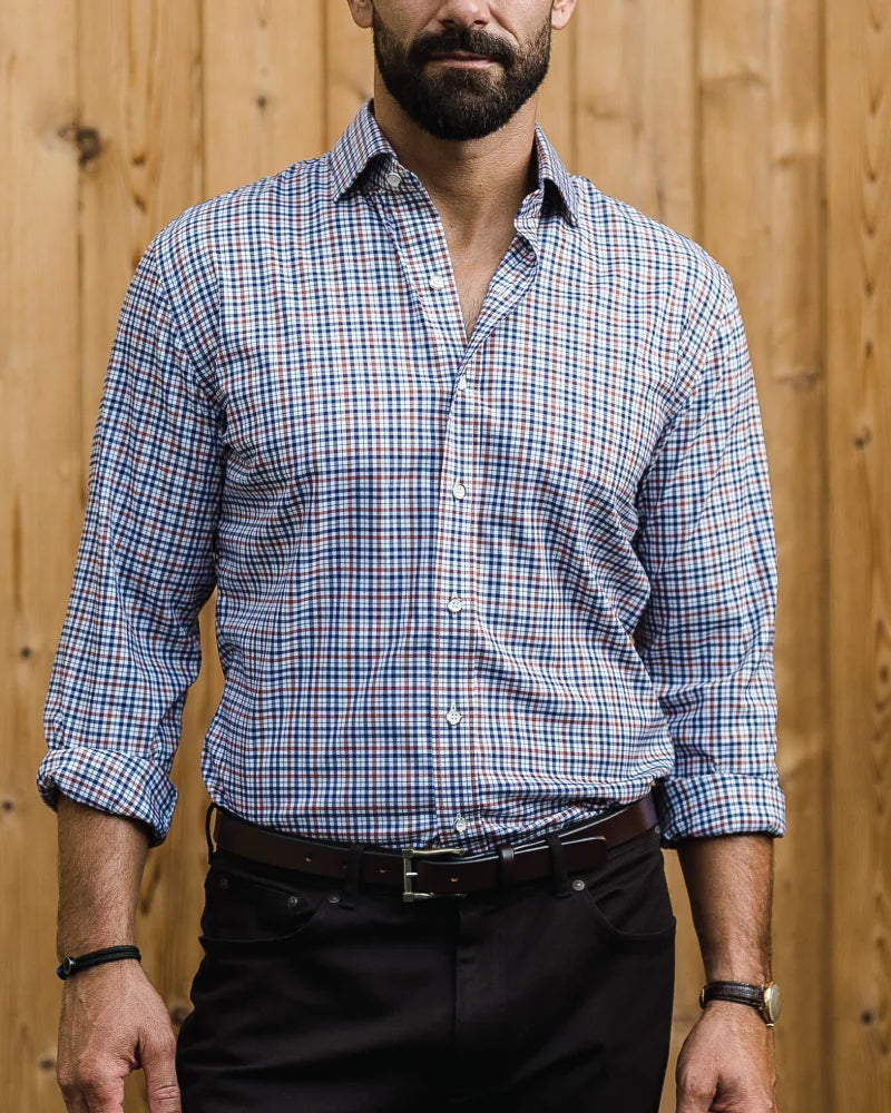 A man wearing a blue, dark brown, and white plaid button-down shirt with black trousers, standing in front of a light brown wooden plank wall.