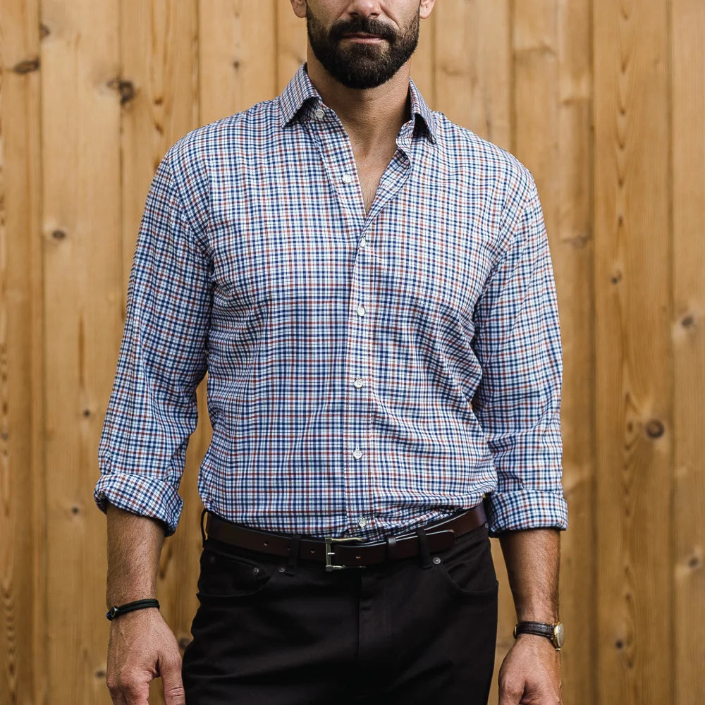 A man wearing a blue, dark brown, and white plaid button-down shirt with black trousers, standing in front of a light brown wooden plank wall.