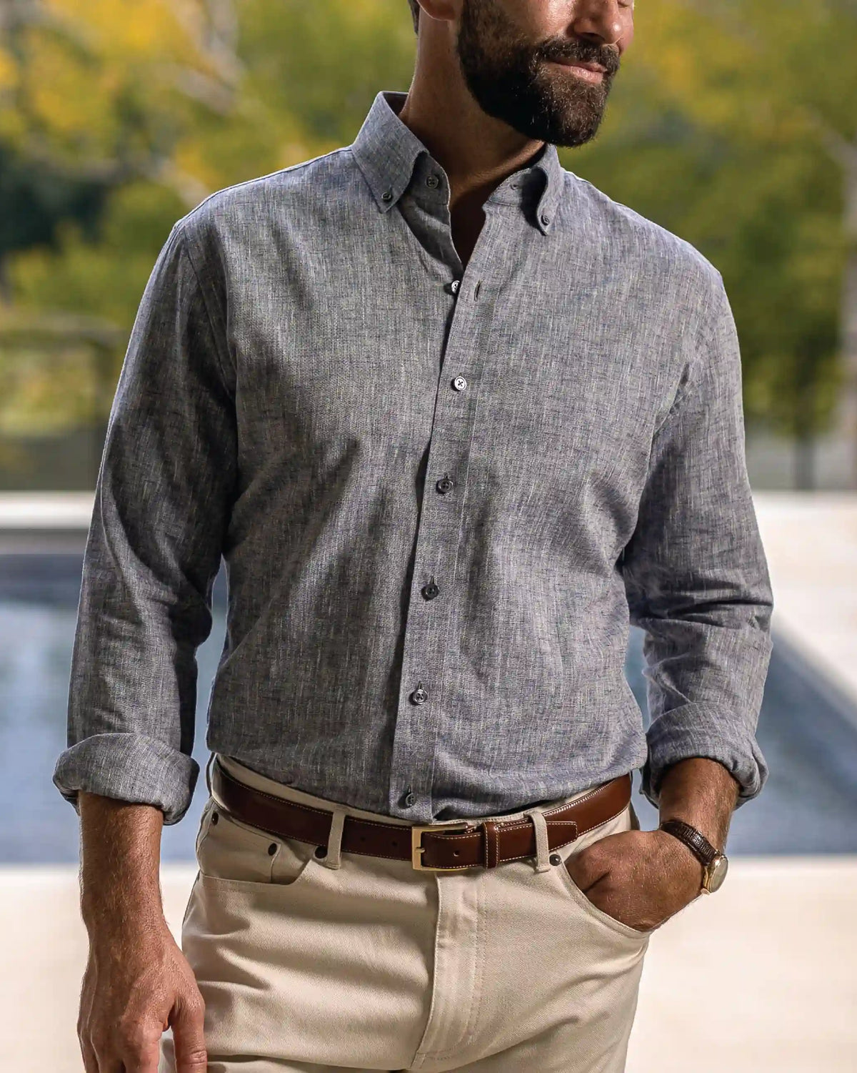 A man wearing a gray long-sleeve linen button-down shirt, paired with beige trousers and a brown belt, standing in front of a blurred outdoor background.