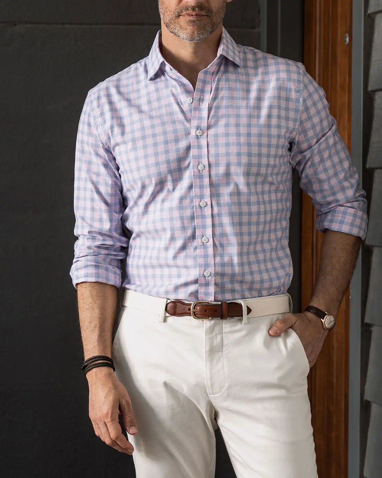 Man wearing a pink and blue checkered performance shirt and white pants standing against a dark wall.